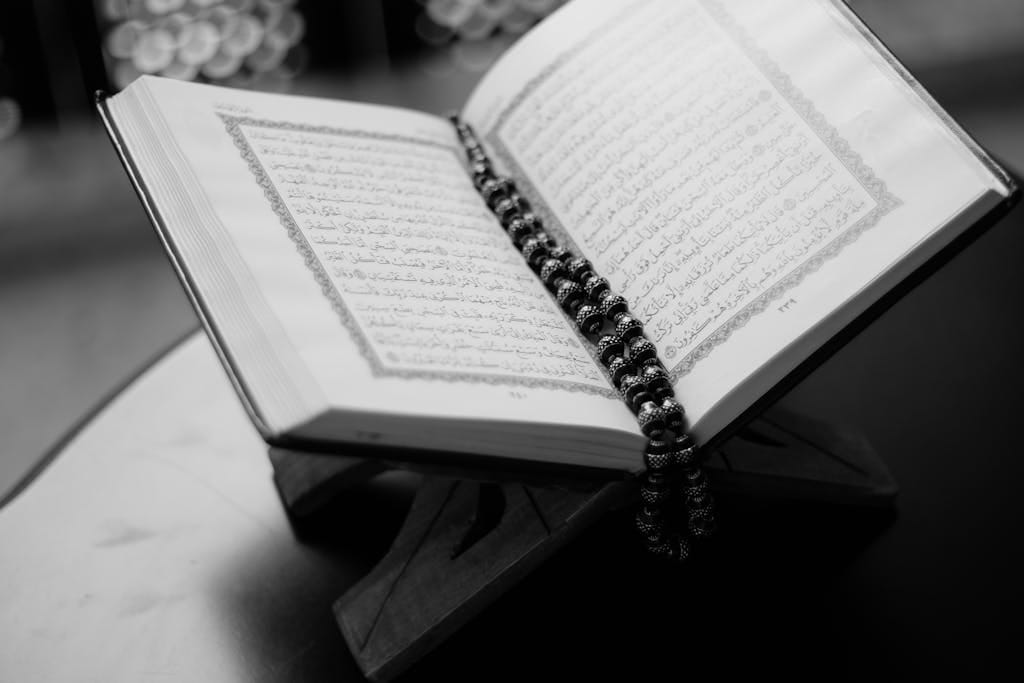 The Importance of Learning the Quran: A Complete Guide to Spiritual Growth 2 A black and white close-up of an open Quran on a wooden stand with prayer beads, symbolizing Islamic faith and devotion.