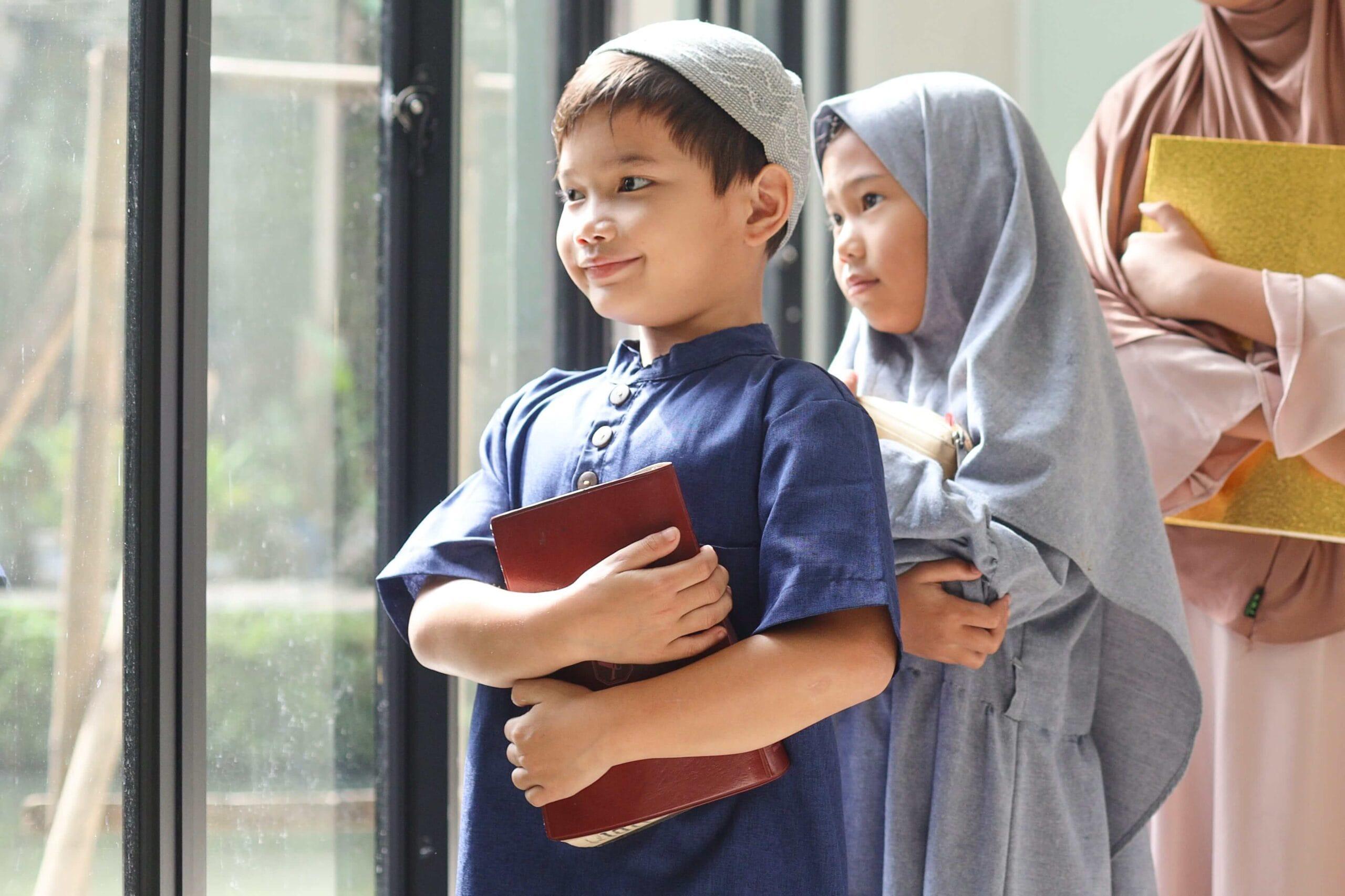 Young Muslim boy with kufi and book standing with two girls in hijabs holding books