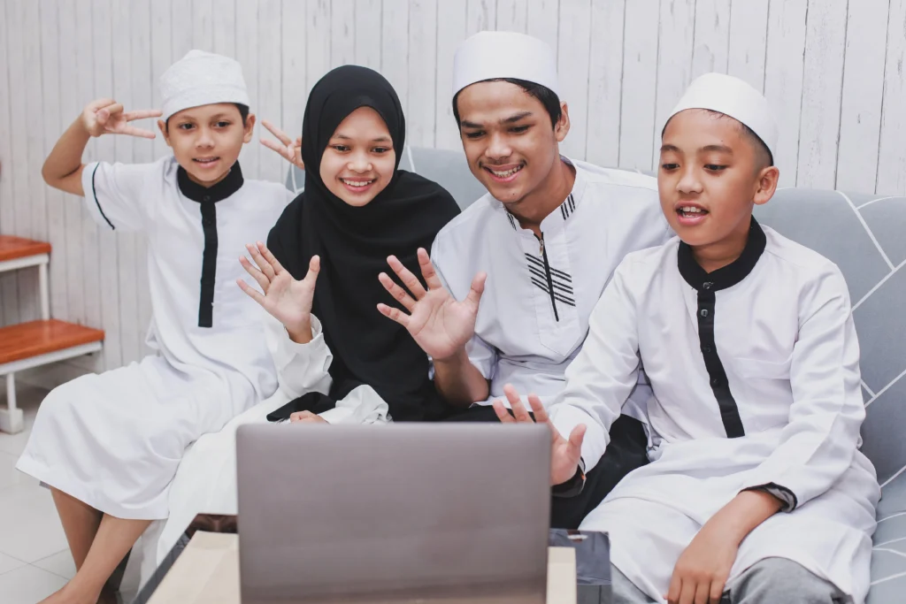Online Quran Memorization Course 2 A group of four Muslim children and teenagers, including a boy wearing a skullcap (kufi) and a girl wearing a hijab, waving at a laptop during a video call.