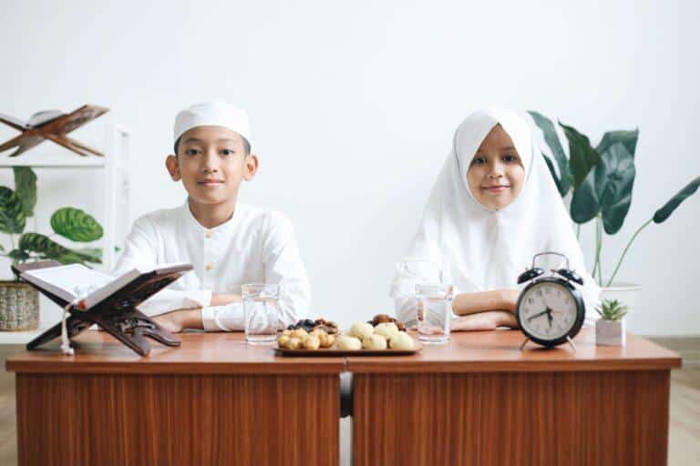 Boy and girl in white Islamic attire at a table with dates, snacks, water, and a clock