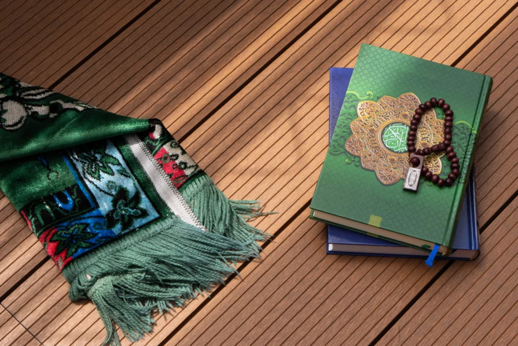 Online Quran Memorization Course 5 A green prayer rug is folded beside a stack of books, with the top one being a green and gold Holy Quran. A string of wooden prayer beads rests on the cover.