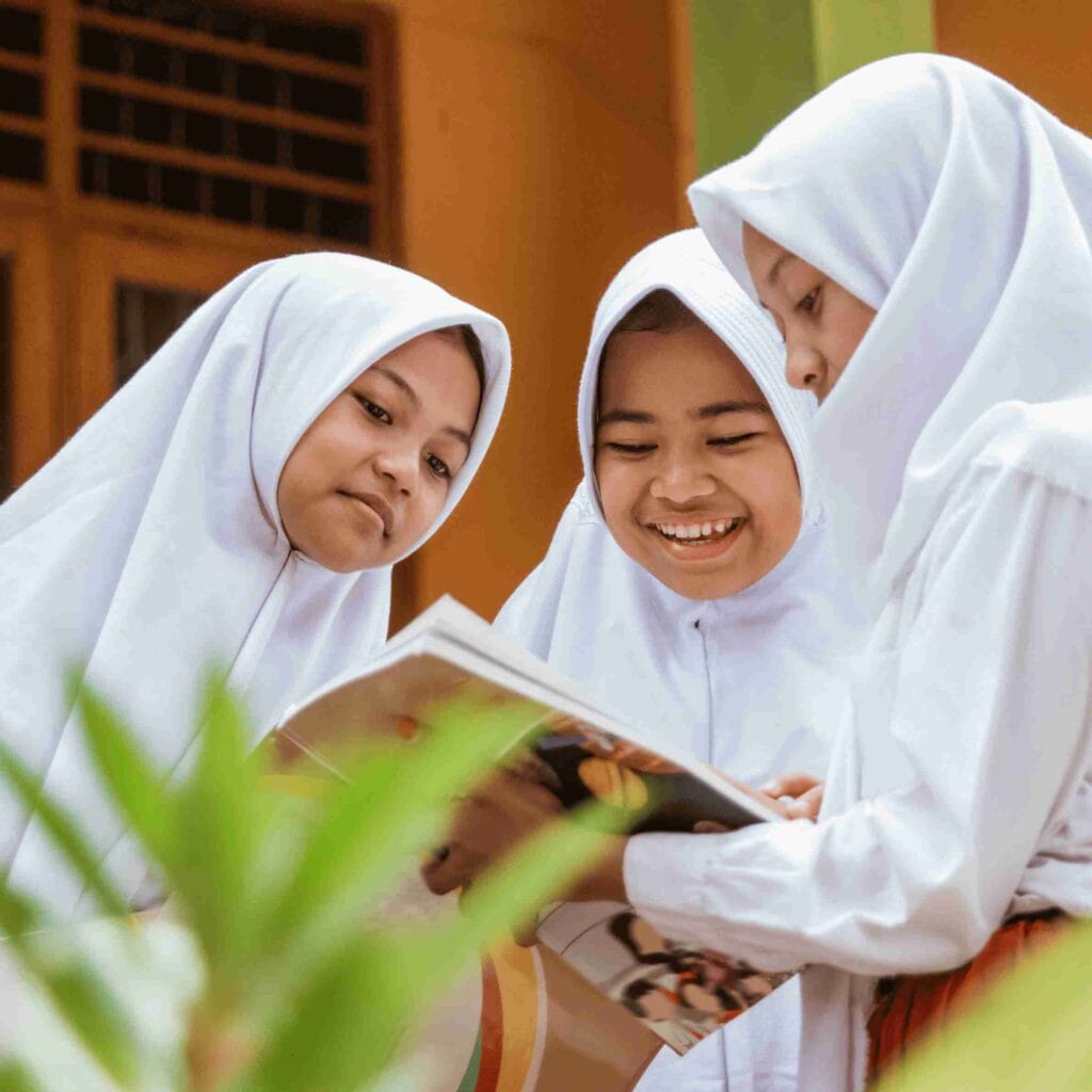 Three Muslim girls in white uniforms and headscarves smiling while reading a book together