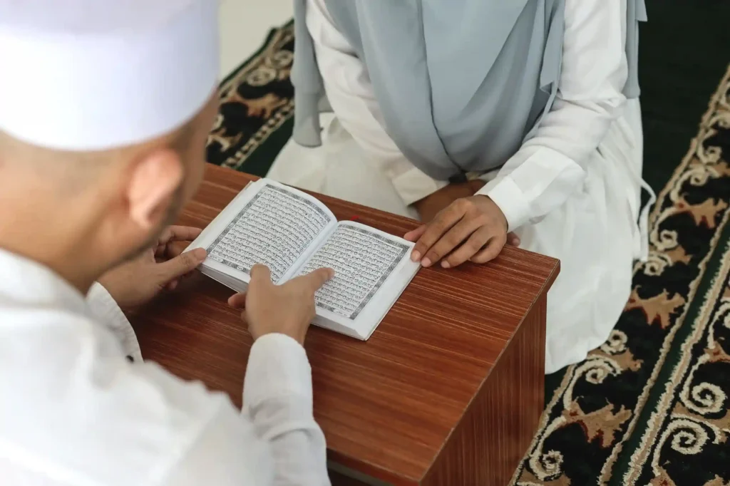 Man and woman in white Islamic attire studying an open Quran together on the floor