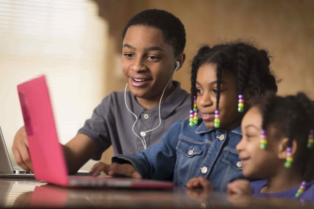 Three children smiling around a pink laptop, with an older boy wearing earphones