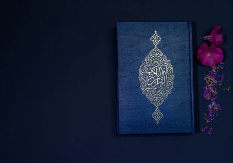 A blue Quran with golden Arabic calligraphy placed on a dark background, accompanied by pink and purple flowers, symbolizing spirituality and devotion.